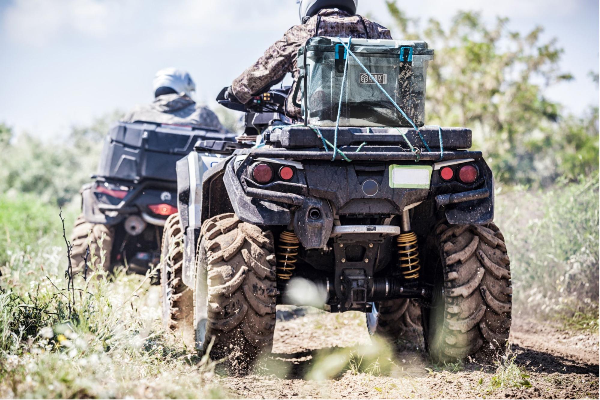 Two people riding an ATV with a QuadraX Cubix Cooler strapped to the back